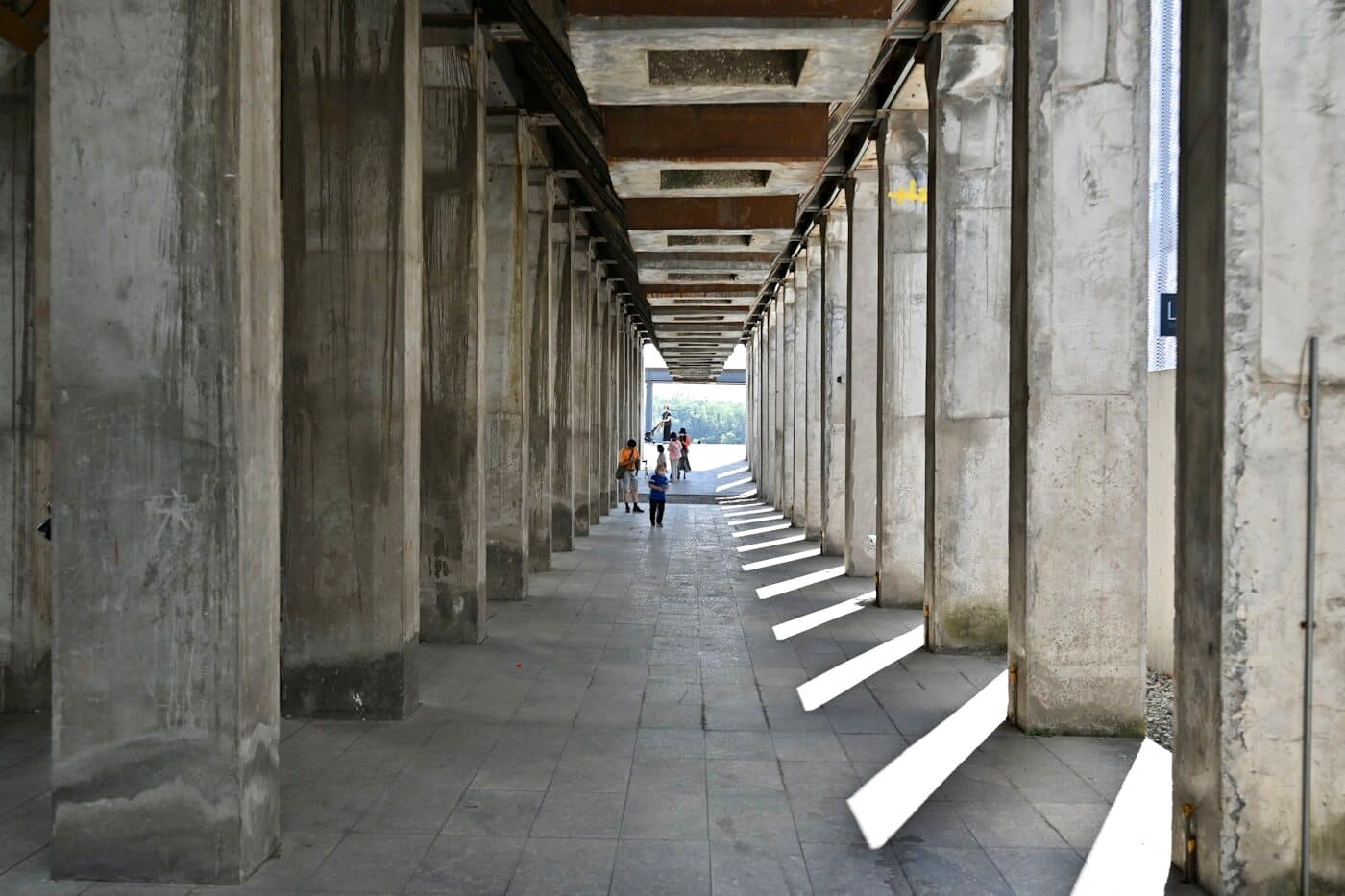 A long colonnade of concrete columns with distant figures walking through the light at its end